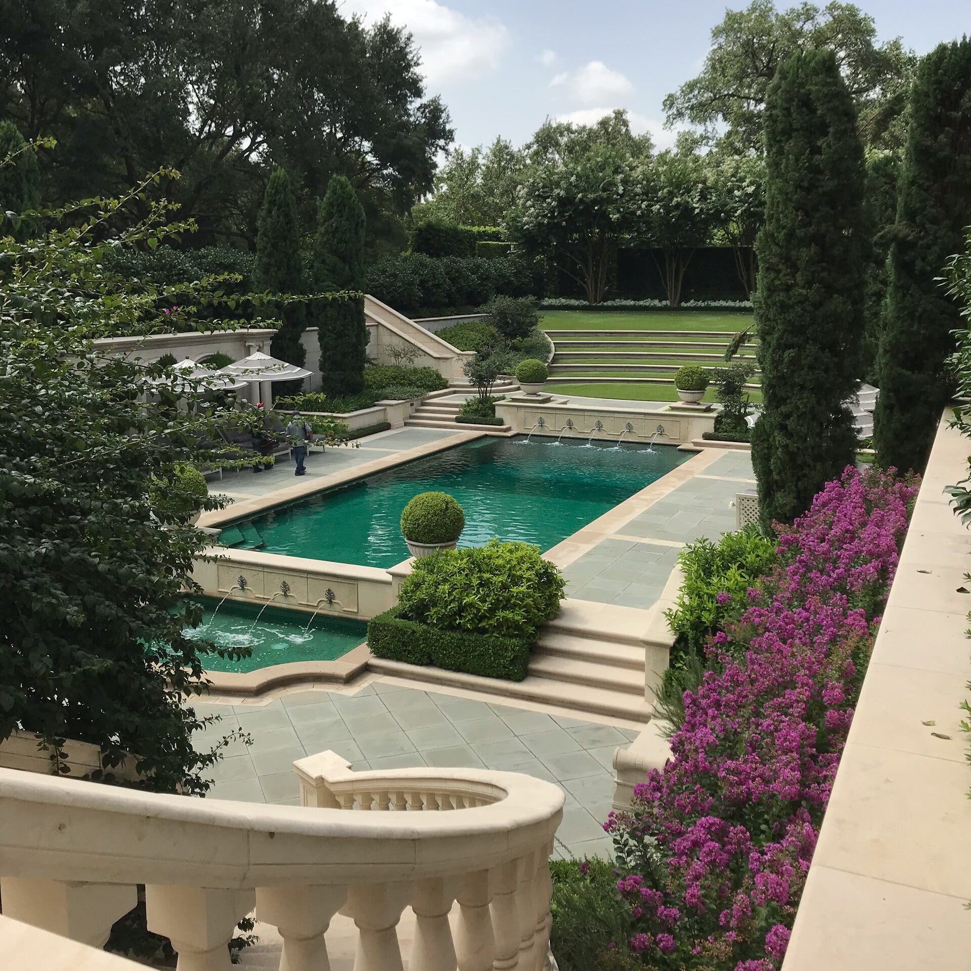 A high-angle view of a luxury rectangular swimming pool with integrated fountains, stone tiered terraces, manicured lawn steps, and vibrant purple flowers.