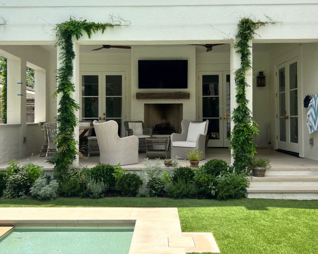 A luxury white covered patio with comfortable wicker seating, an outdoor fireplace, a wall-mounted TV, and a pool in the foreground.