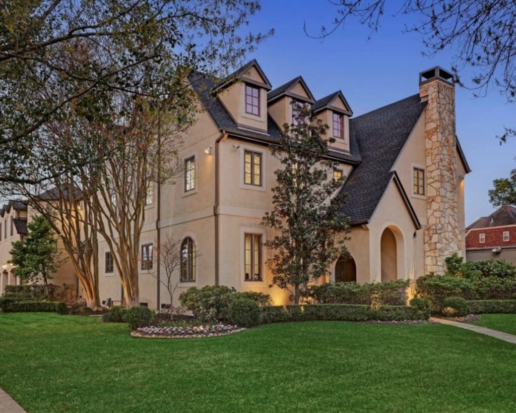 The original front view of a three-story beige stucco home with a high-pitched roof and a large stone chimney before its renovation.